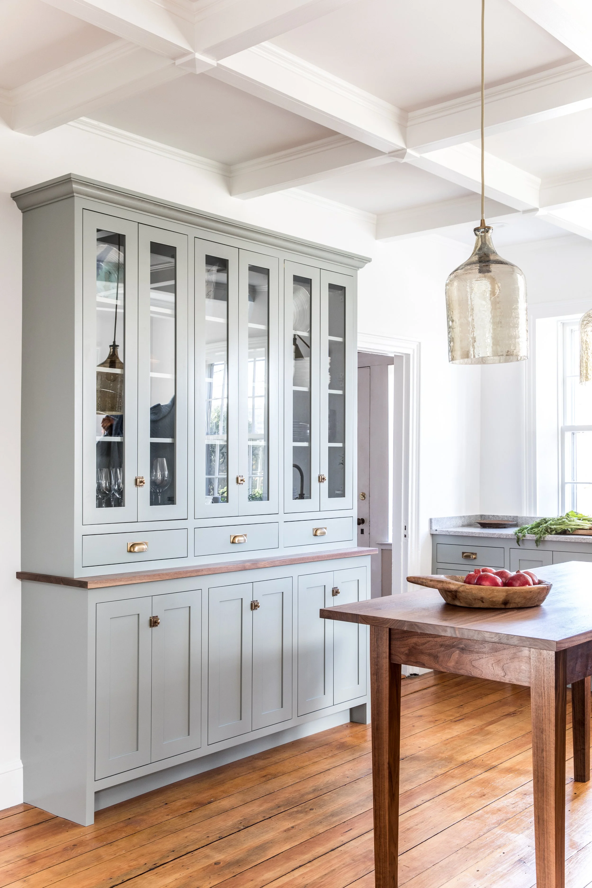 A light gray kitchen cabinet with glass doors, gold hardware, and a wooden countertop, with a wooden table and bowl of apples nearby, in a bright room with wooden floors and a large window.