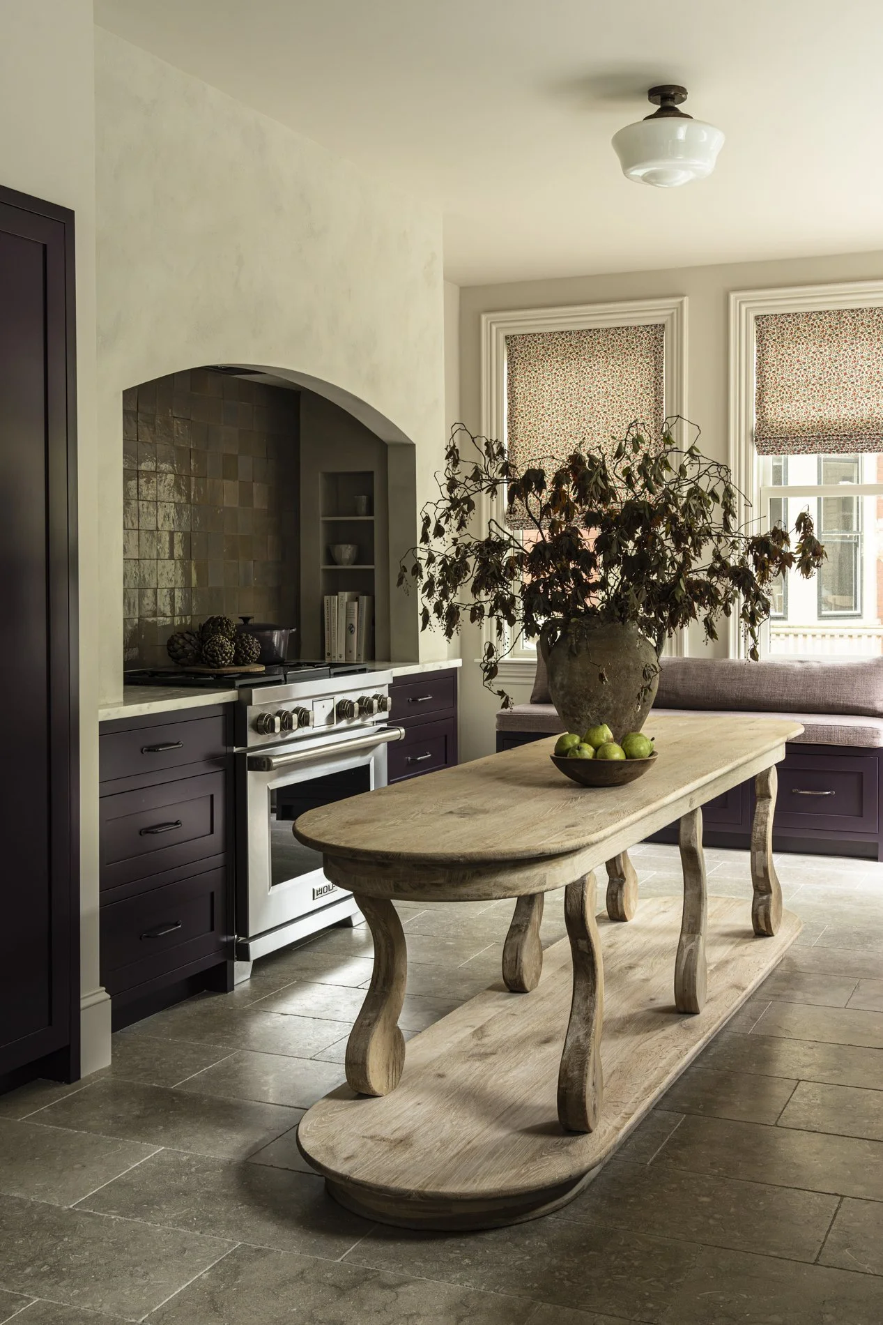 A rustic kitchen with purple cabinetry, a stainless steel stove, and a wooden table with curved legs and a large vase with dried branches, set near two windows with patterned blinds.