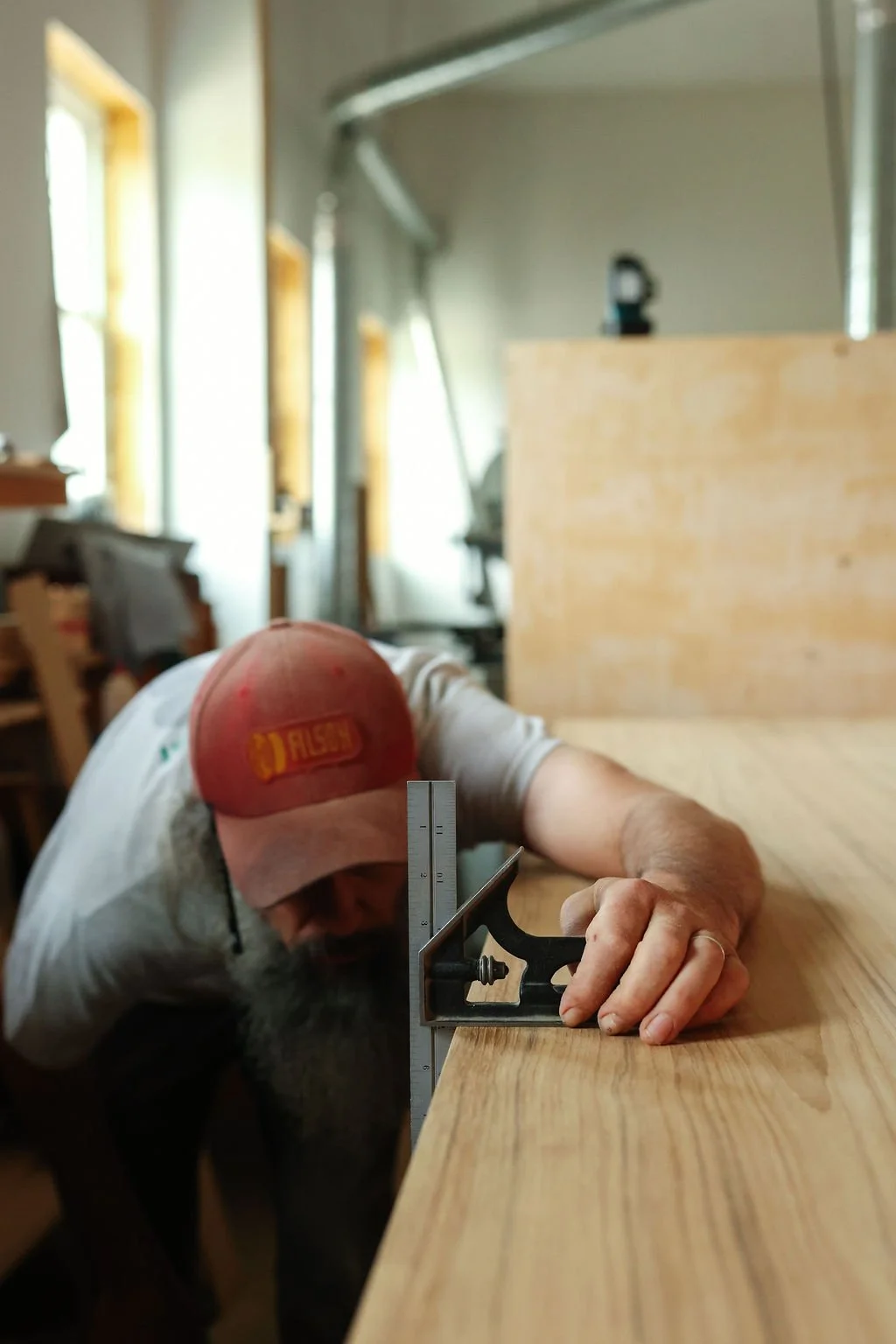 A man working on a woodworking project, using a square to measure a piece of wood in a workshop with natural light from windows.