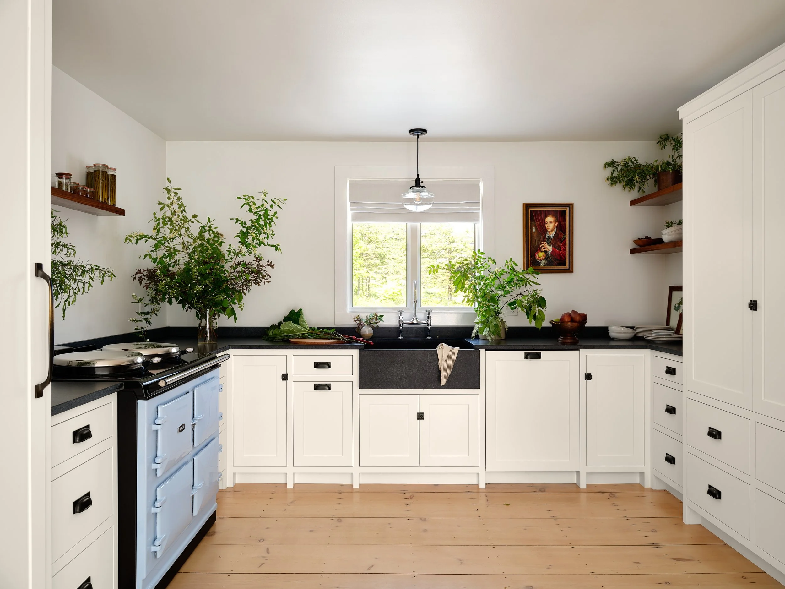 Kitchen with white cabinets, black countertop, black farmhouse sink, window with blinds, plants, and a blue stove, wooden floors, and open shelves with jars and dishes.