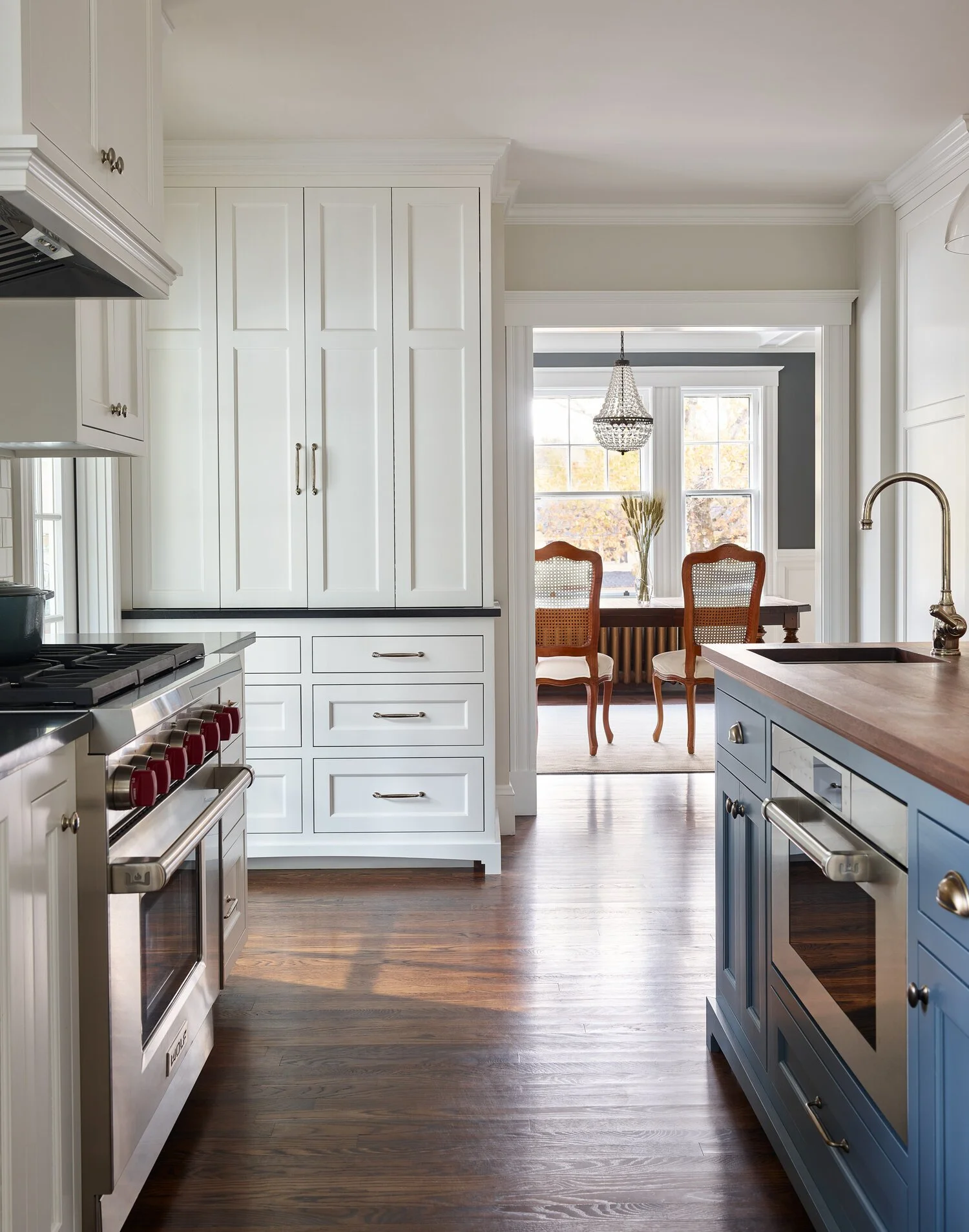 A bright kitchen with white cabinetry, a stainless steel oven with red knobs, and a blue kitchen island with a wooden countertop. In the background, a dining area with a wooden table, two chairs, and a chandelier is visible through a doorway. Large w