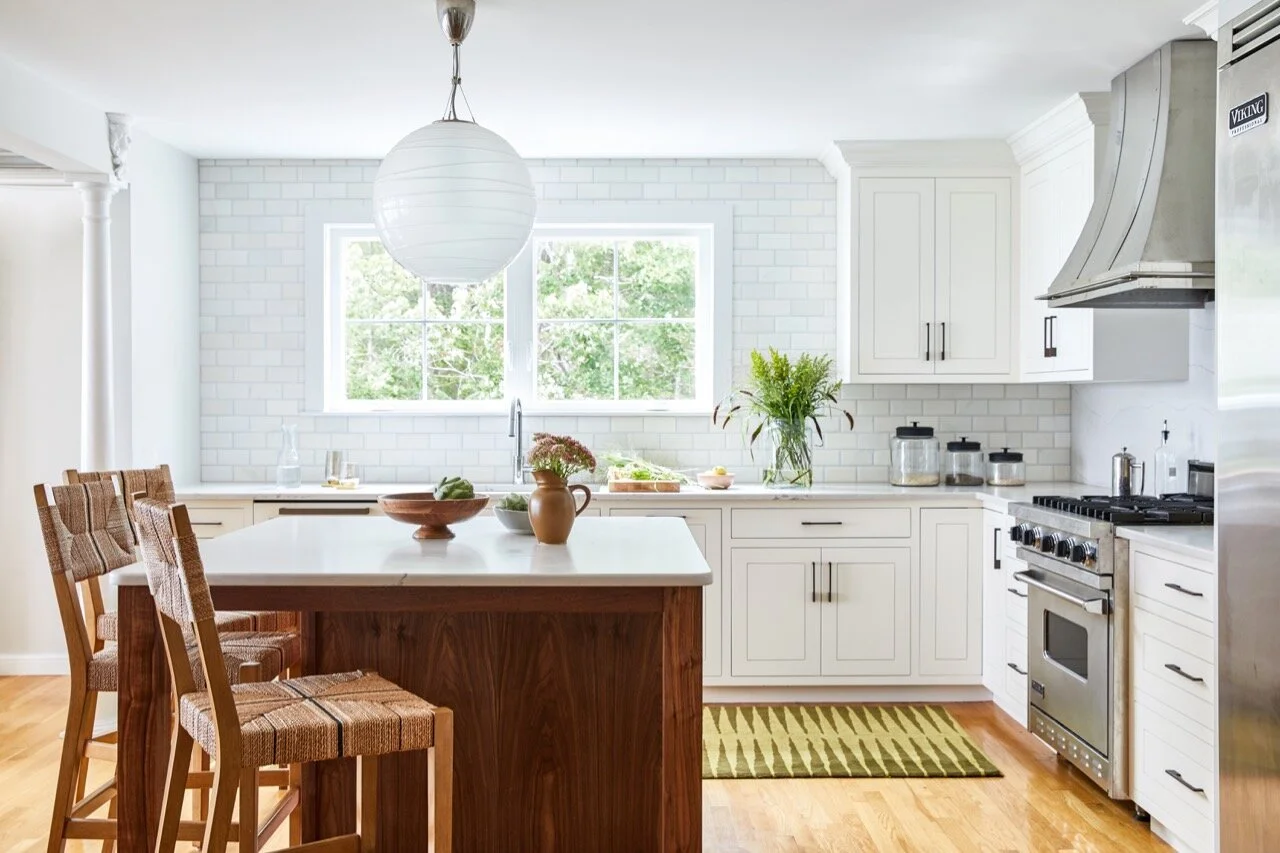 Bright kitchen with white cabinets, stainless steel stove, and a wooden island with three chairs, decorated with vases of flowers and greenery.