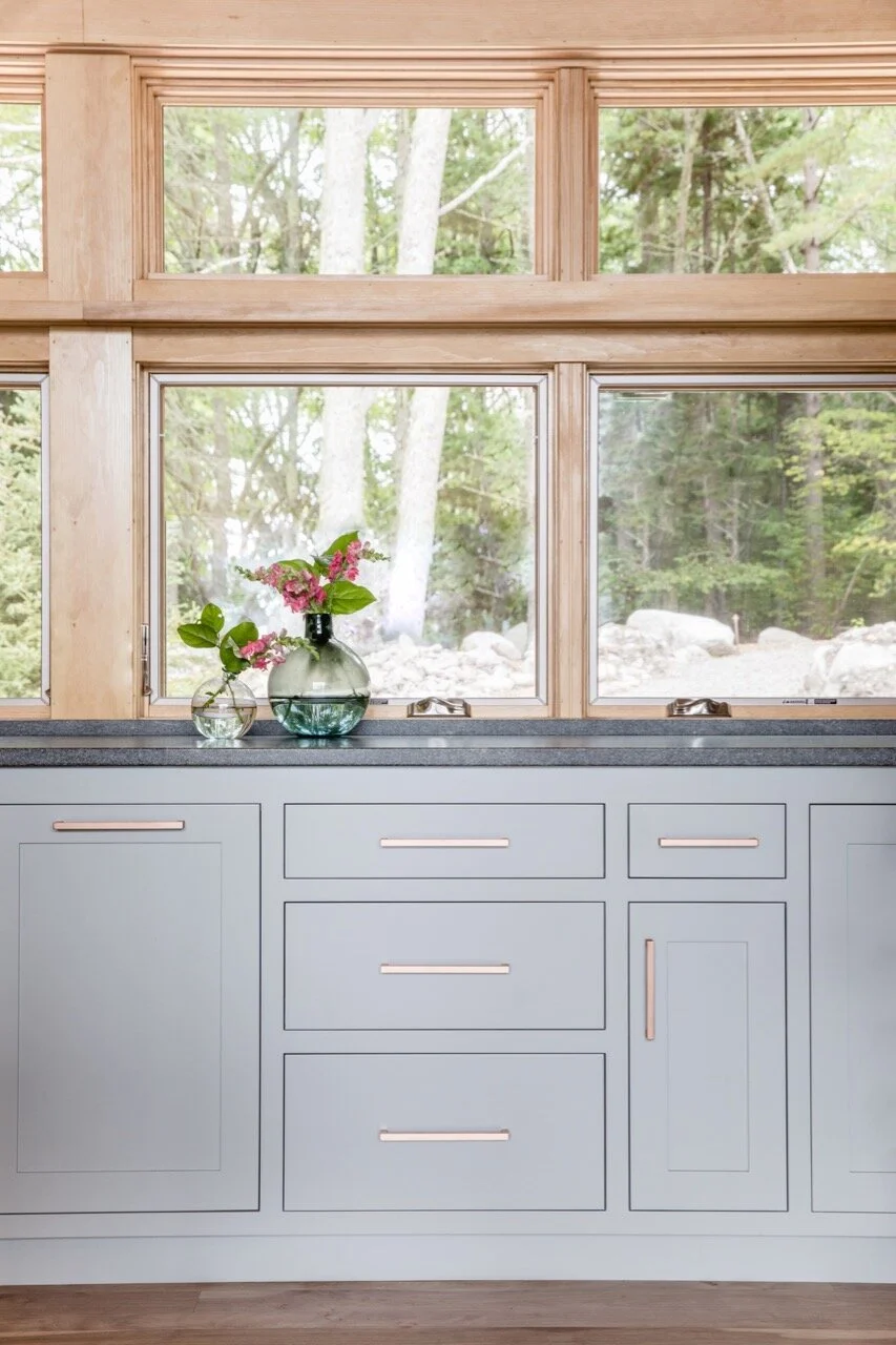 Kitchen window with wooden frame, light blue cabinets, and decorative vases with flowers on the black countertop, overlooking a wooded area outside.