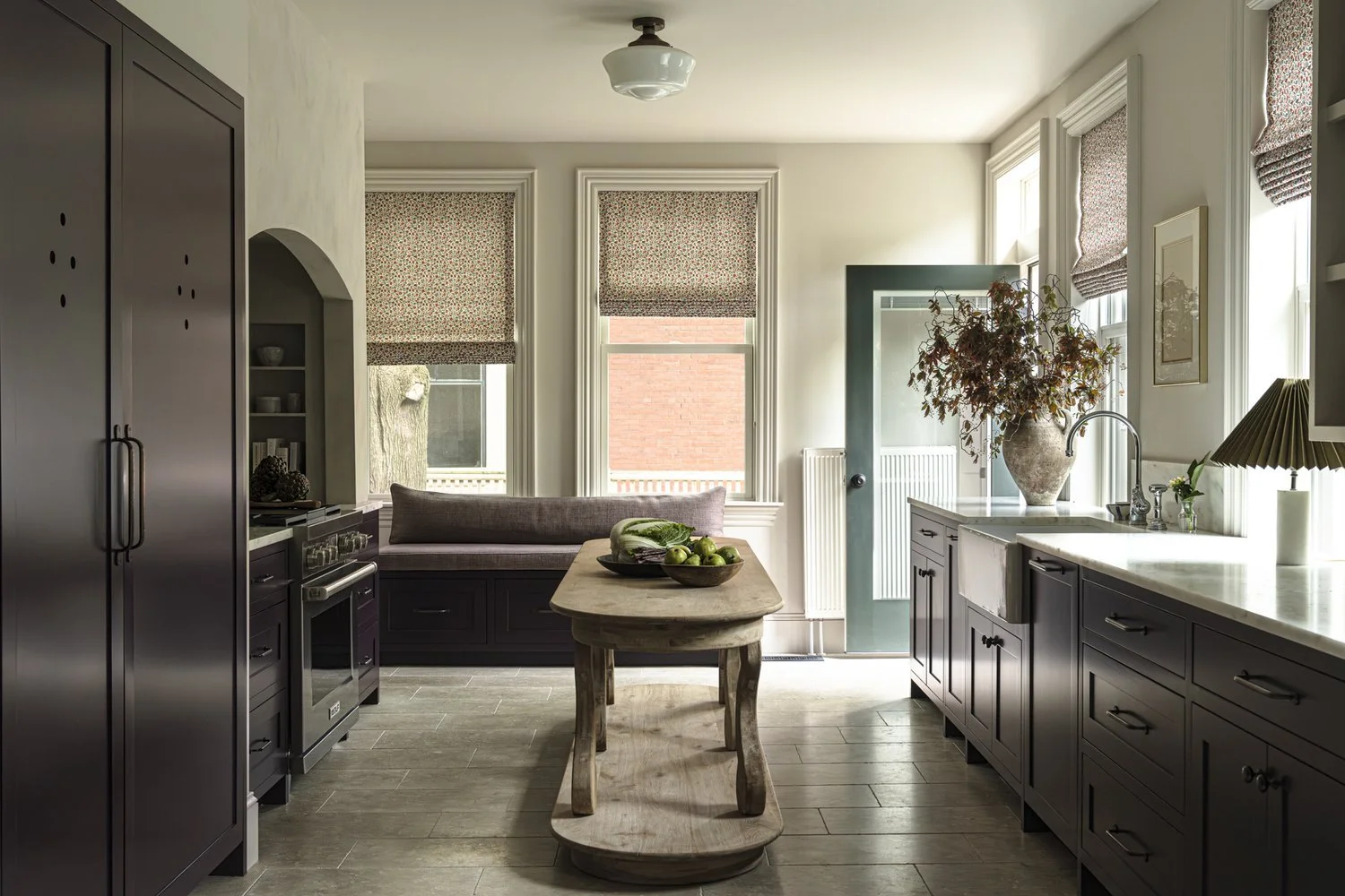 Bright kitchen with dark cabinets, large windows with patterned Roman shades, a wooden table with green apples, and a built-in window seat with a cushion.