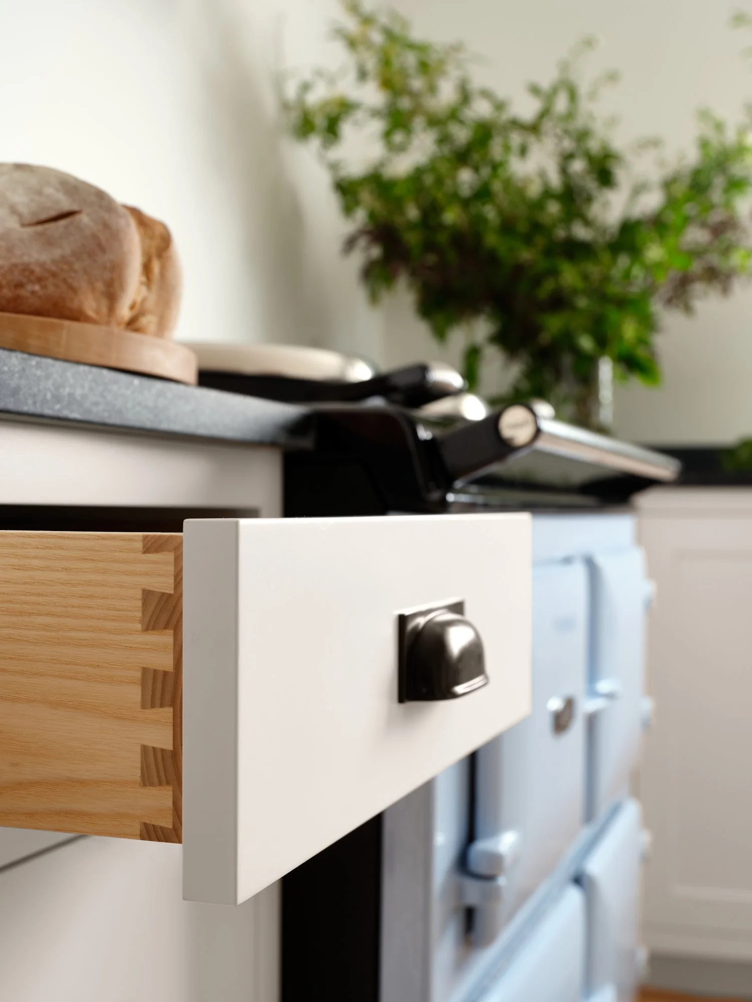 Dovetail detail on kitchen drawer with a black handle, wooden cabinet edge, and blurred kitchen countertop with a loaf of bread, pots, and a plant in the background.