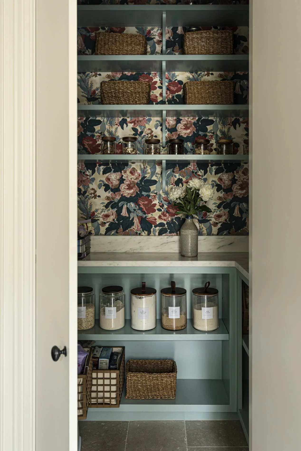 A pantry with teal shelves, floral wallpaper, glass jars of grains and sugar, wicker baskets, and a vase with white flowers.
