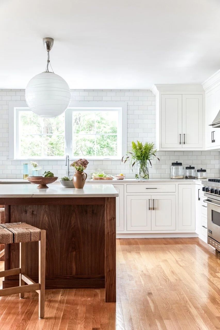 Bright white kitchen with white cabinets, large window, wooden grain-matched walnut island with striped stool, hardwood floor, pendant light, and decorative plants.