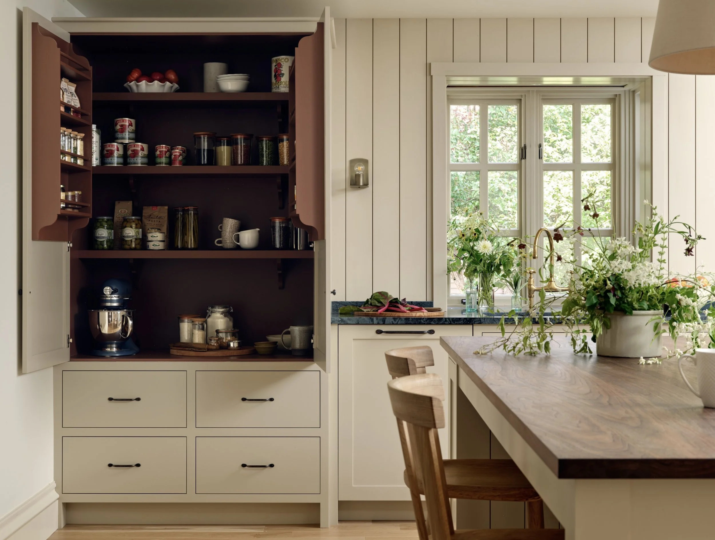 A kitchen with an open cabinet filled with spices, jars, and cups, next to a window with green plants, a wooden table with a flower arrangement, and kitchen utensils.