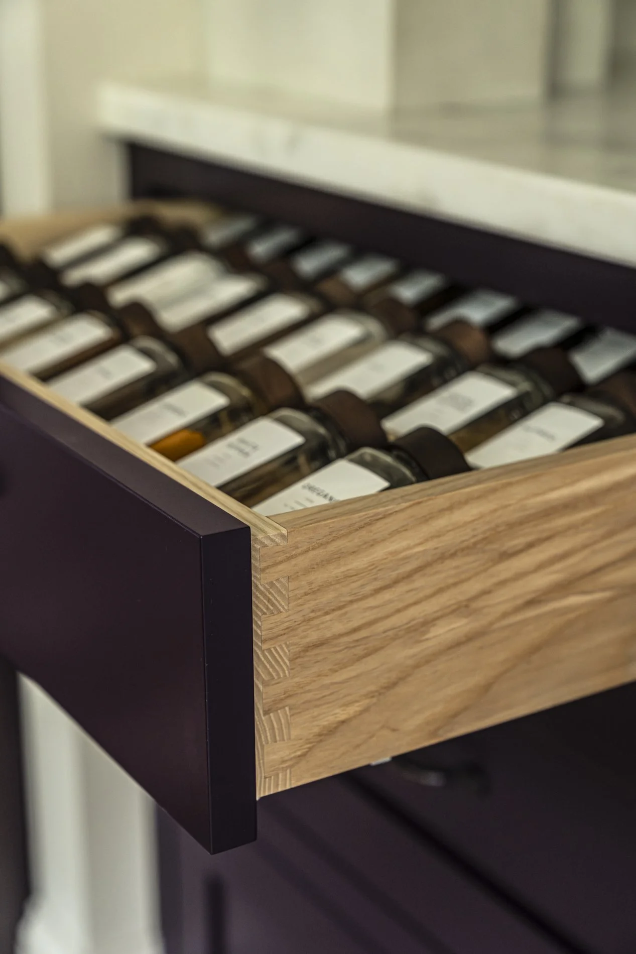 A wooden drawer filled with glass jars, each labeled with white tags, partially pulled out from a purple cabinet.