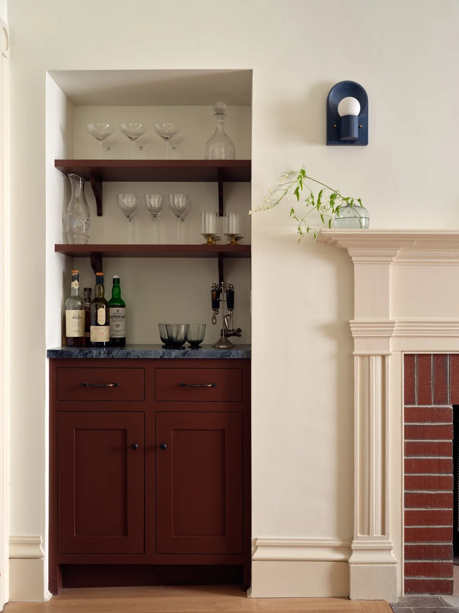A built-in wooden liquor cabinet with glassware on shelves, bottles of alcohol, and decorative items, adjacent to a white fireplace with a small vase and a wall-mounted light fixture.