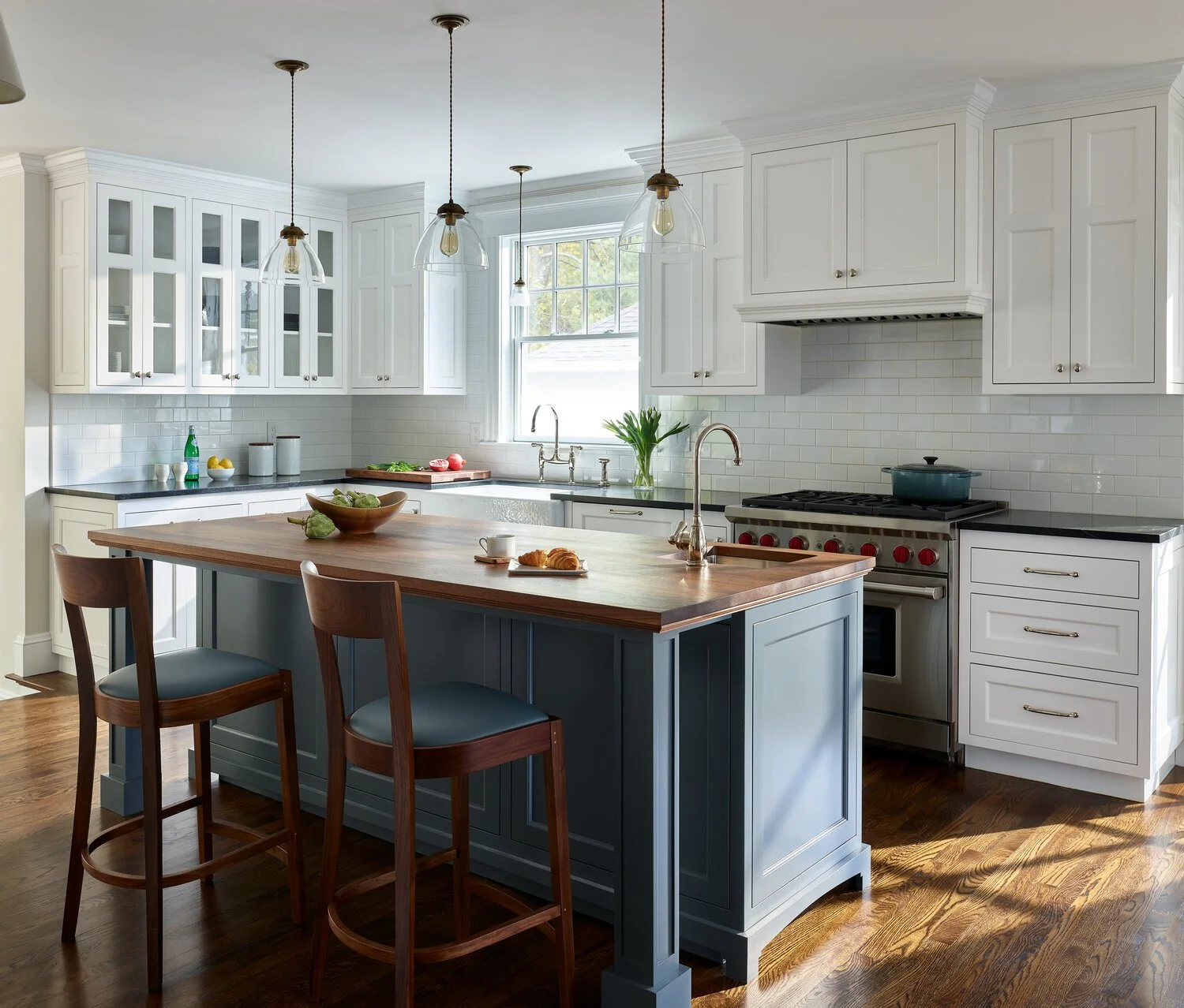 A modern kitchen with white cabinets, a wooden island, and hanging pendant lights, with a window over the sink and hardwood flooring.