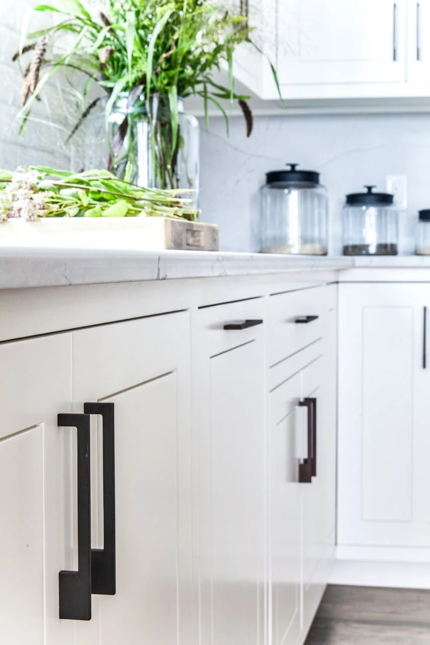 White kitchen cabinets with black handles, a marble countertop, and glass jars with black lids in the background, along with a potted plant and herbs on the countertop.