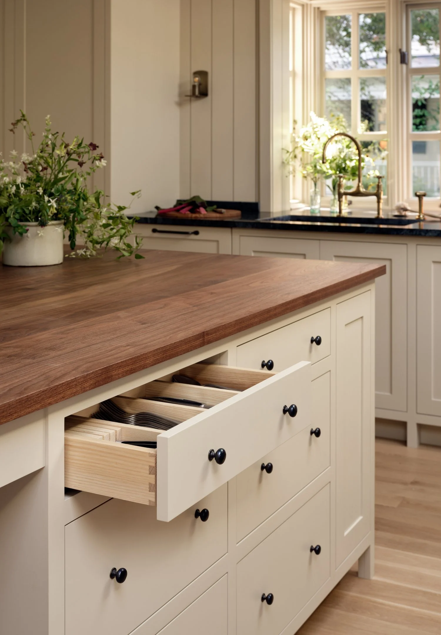 Kitchen with cream cabinets, black knobs, wooden countertop, open drawer with utensils, window with plants, and a vase of white flowers.