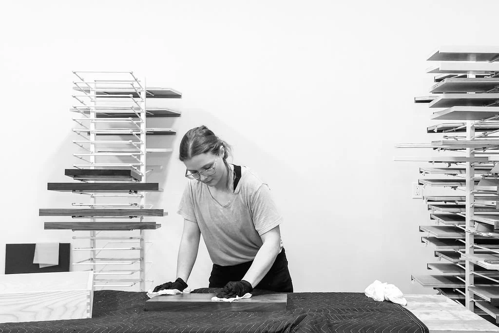 A woman wearing glasses, gloves, and casual clothes is working on a woodworking project at a table, with organized shelves of materials behind her.