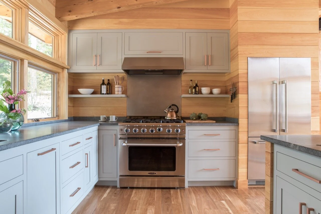 Modern kitchen with light gray cabinets, stainless steel stove and refrigerator, wooden walls, and large windows with a view of trees outside.