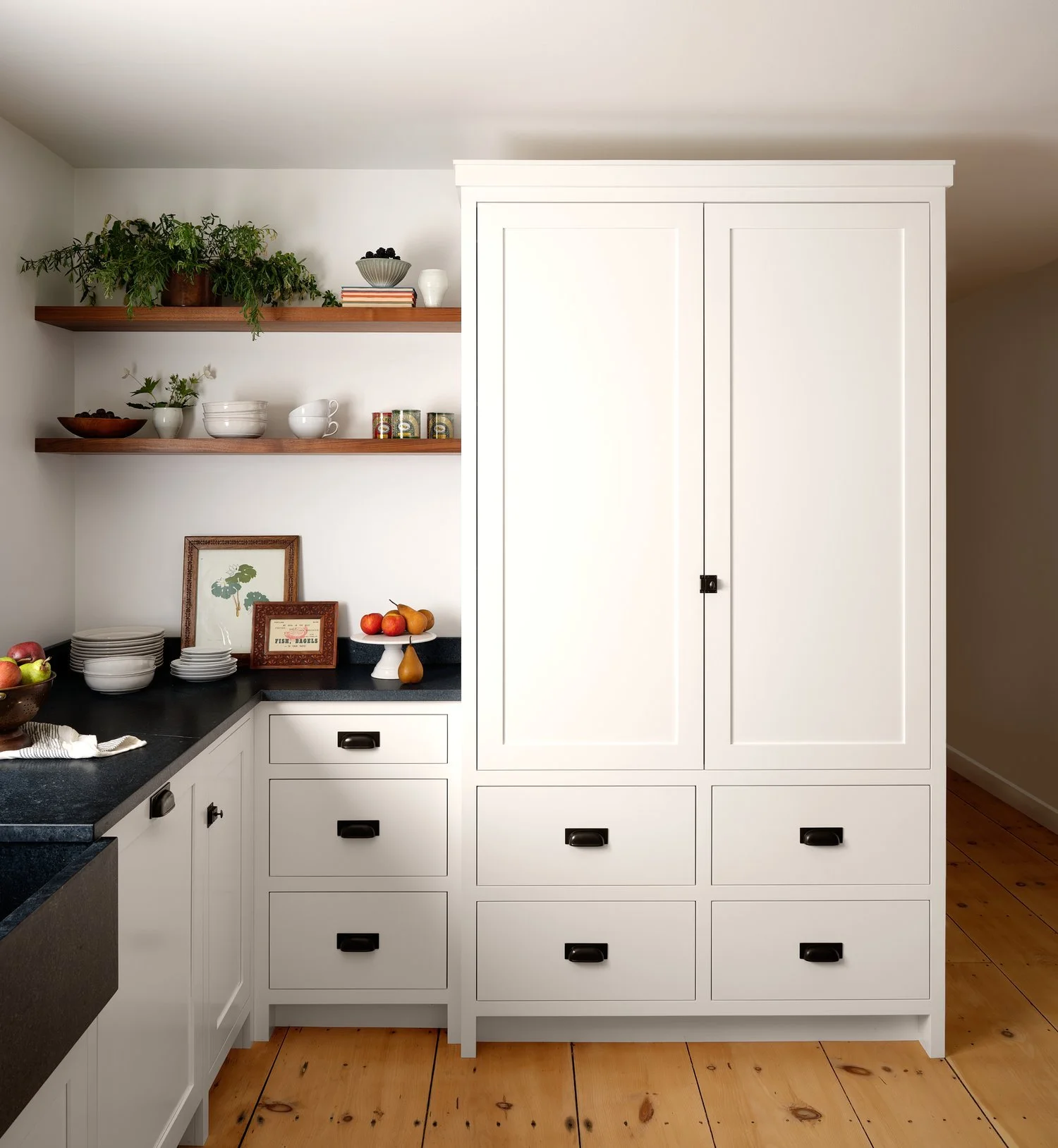 A white kitchen cabinet with multiple drawers and a tall cupboard, black handles, open wooden shelves with bowls, plates, books, and a potted plant, a black countertop with bowls of fruit and decorative items, and a wooden floor.