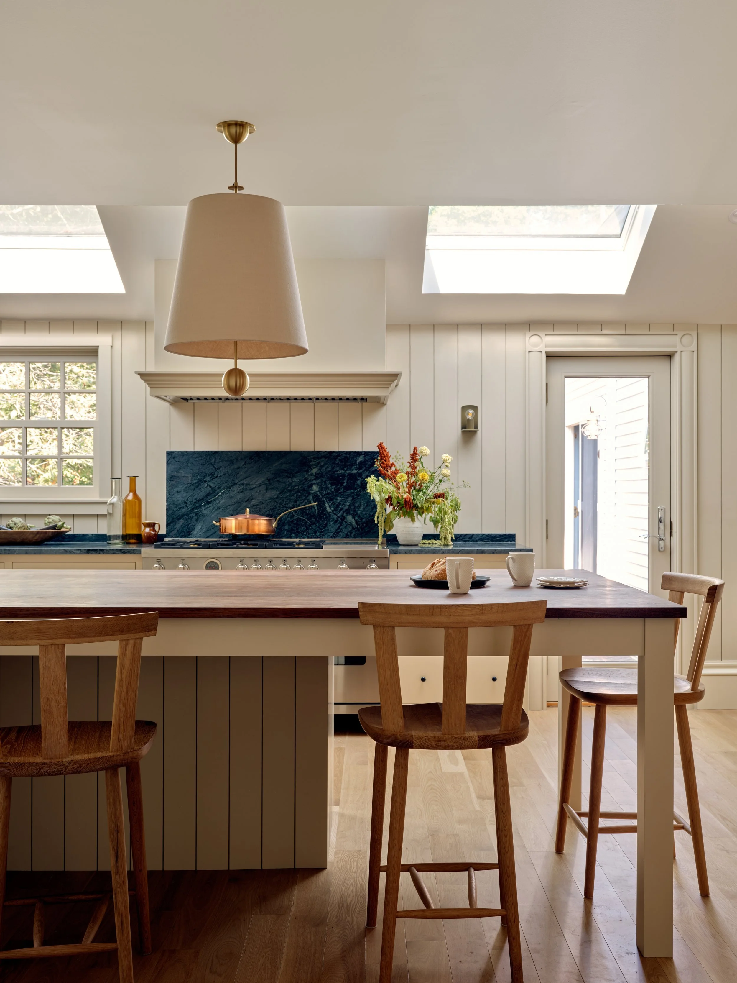 Kitchen with white panel walls, a wooden island with chairs, a copper pan on a stove beneath a vent hood, a vase with flowers, and natural light from skylights and a door.