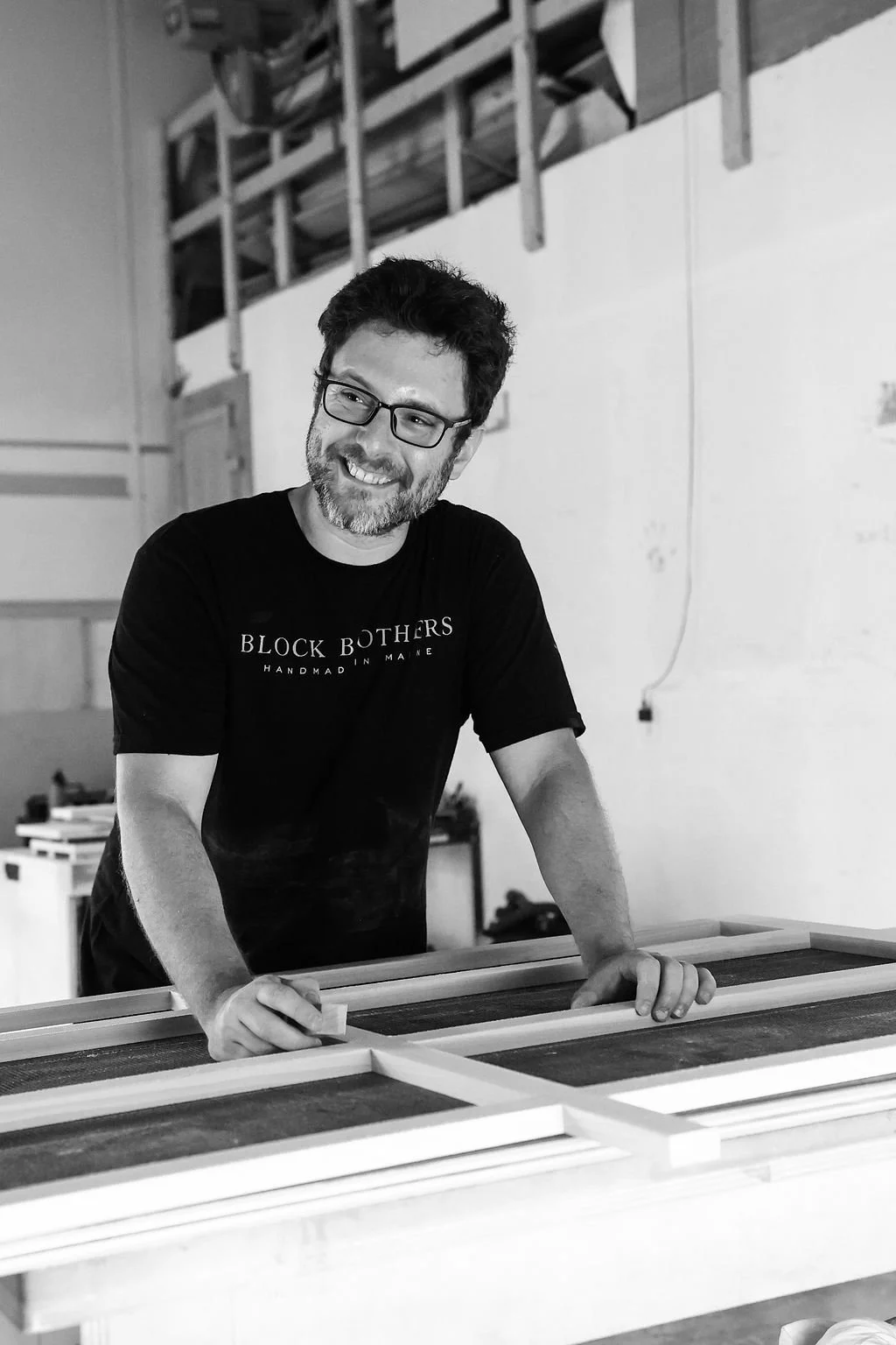 A man with glasses and a beard smiling while working on a woodworking project in a workshop, wearing a T-shirt that says 'Block Brothers Handmade in Maine'.
