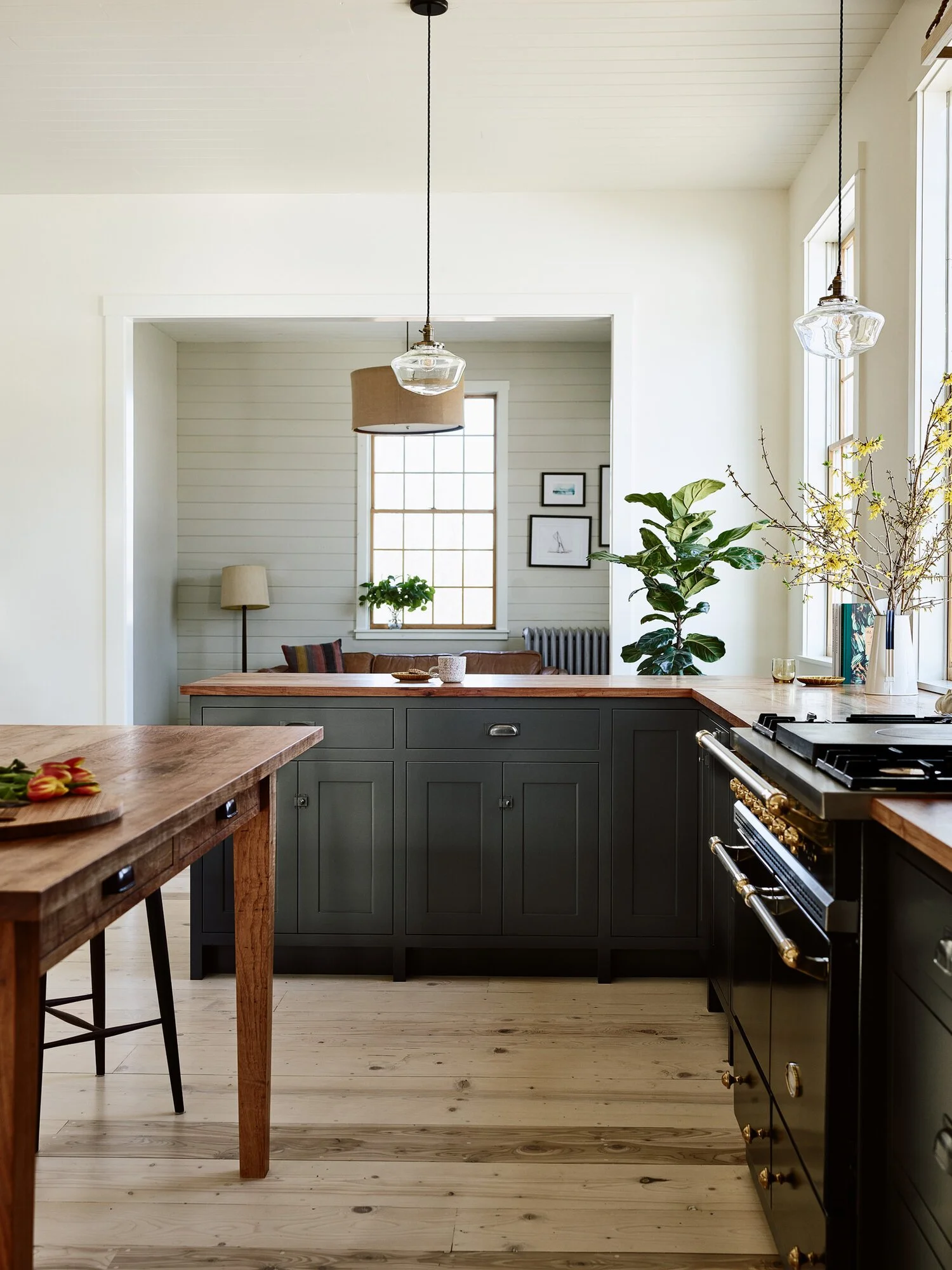 Modern kitchen with black cabinetry, wooden countertops, a stove, and hanging pendant lights, adjacent to a dining area with a wooden table and natural light from large windows, decorated with plants and vases.
