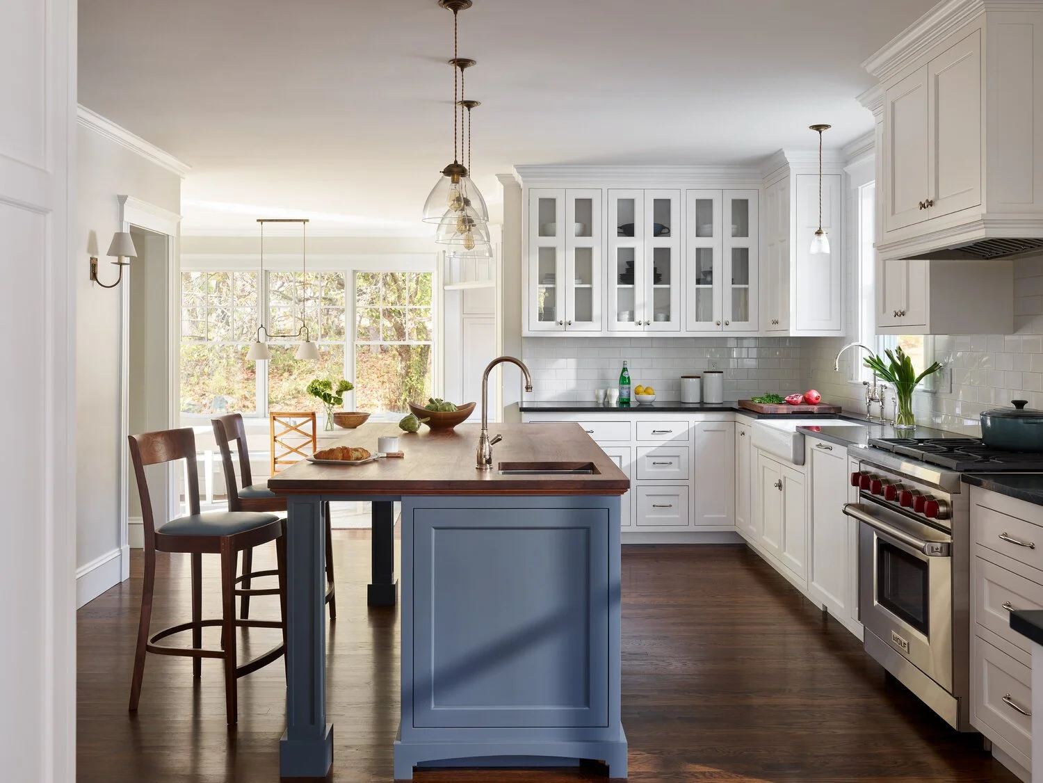 Bright kitchen with white cabinets, a blue kitchen island, and a dining area with large windows showing trees outside.