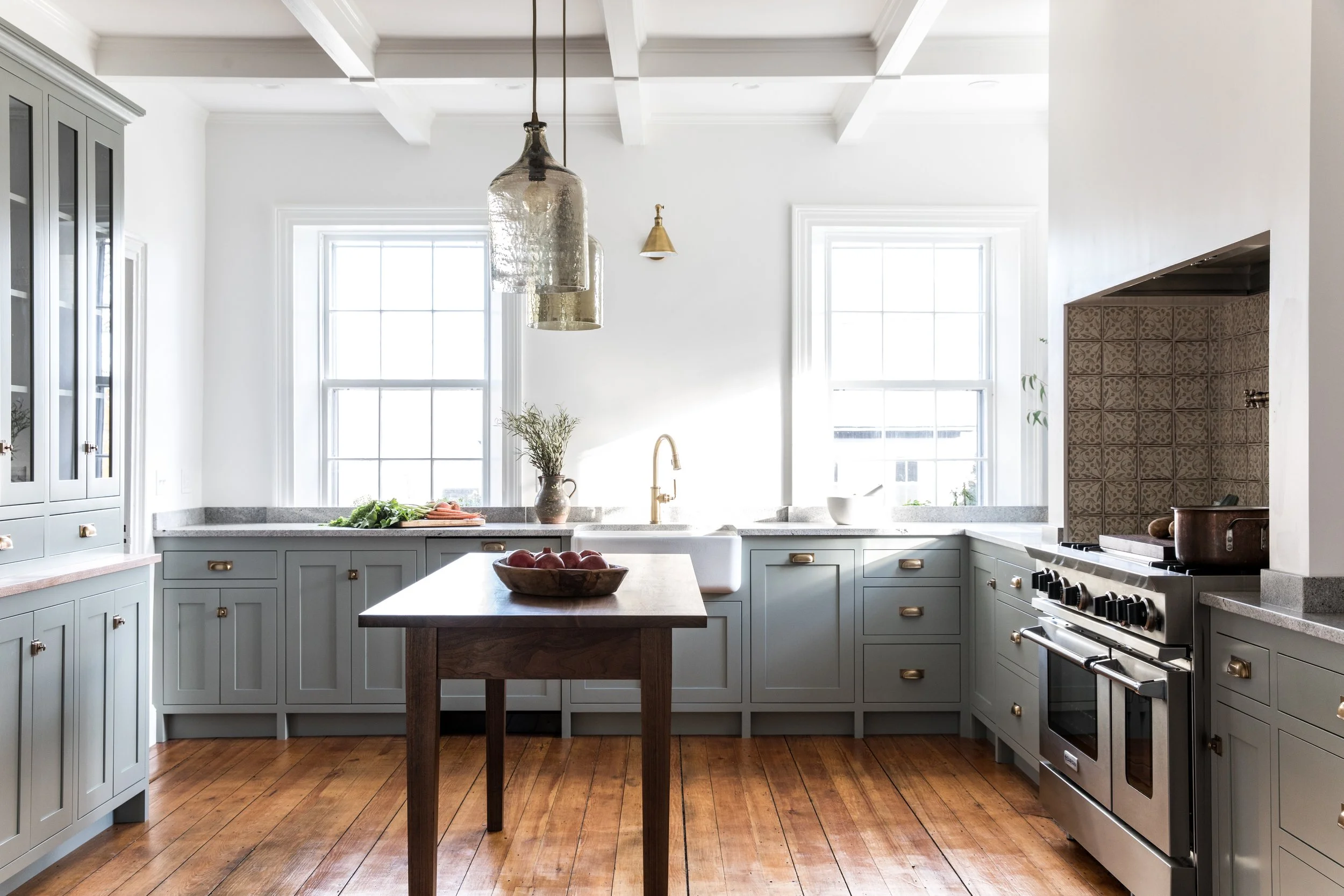 Bright kitchen with light gray cabinets, a central wooden island with a bowl of apples, two large windows, a white farmhouse sink, brass fixtures, pendant lights, and a stainless steel stove, with wood flooring.
