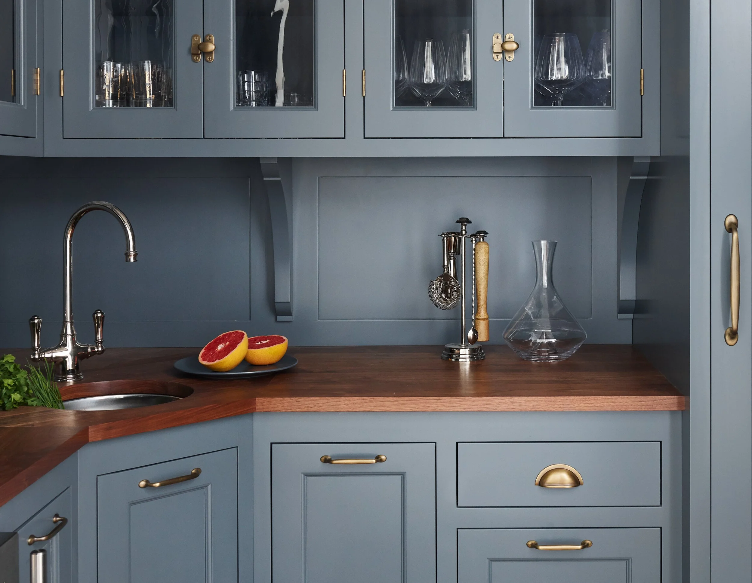 Butler's Pantry/Scullery with down pipe cabinets, wooden countertop, a sink, two halves of grapefruit on a plate, some greenery, and glassware on shelves.
