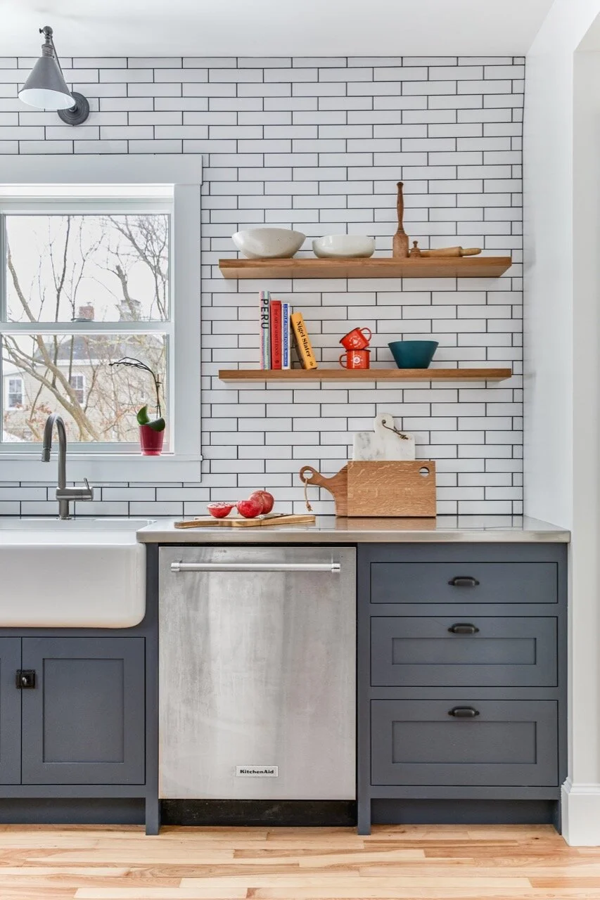 Kitchen with white subway tile backsplash, black faucet, window with a potted plant, open wooden shelves with bowls, books, and decor, gray cabinetry, stainless steel dishwasher, and wooden countertop with apples and a cutting board.