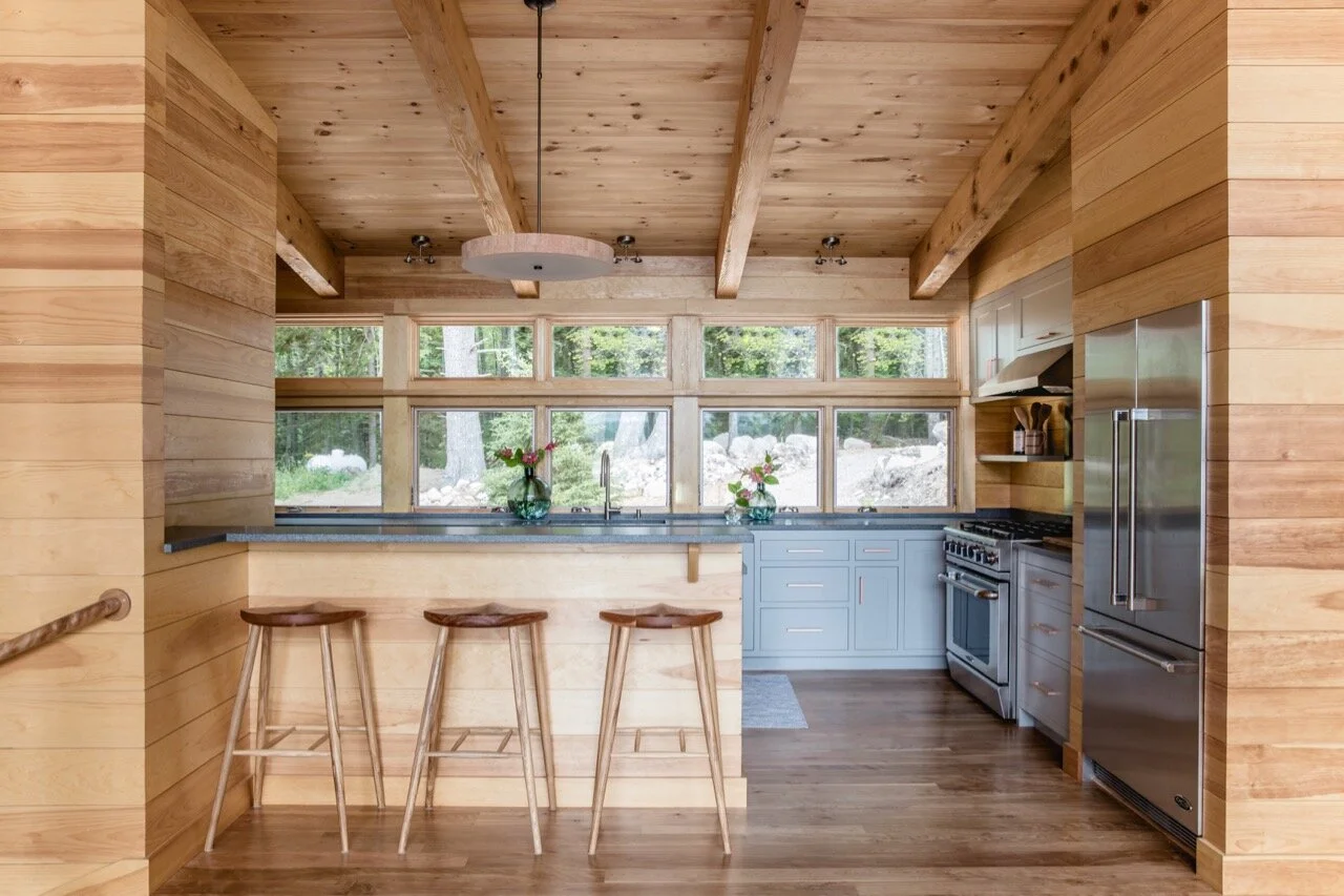 Rustic kitchen with wooden walls and ceiling, blue cabinets, stainless steel refrigerator, stove, and large windows overlooking a wooded area, with three wooden bar stools at the counter.