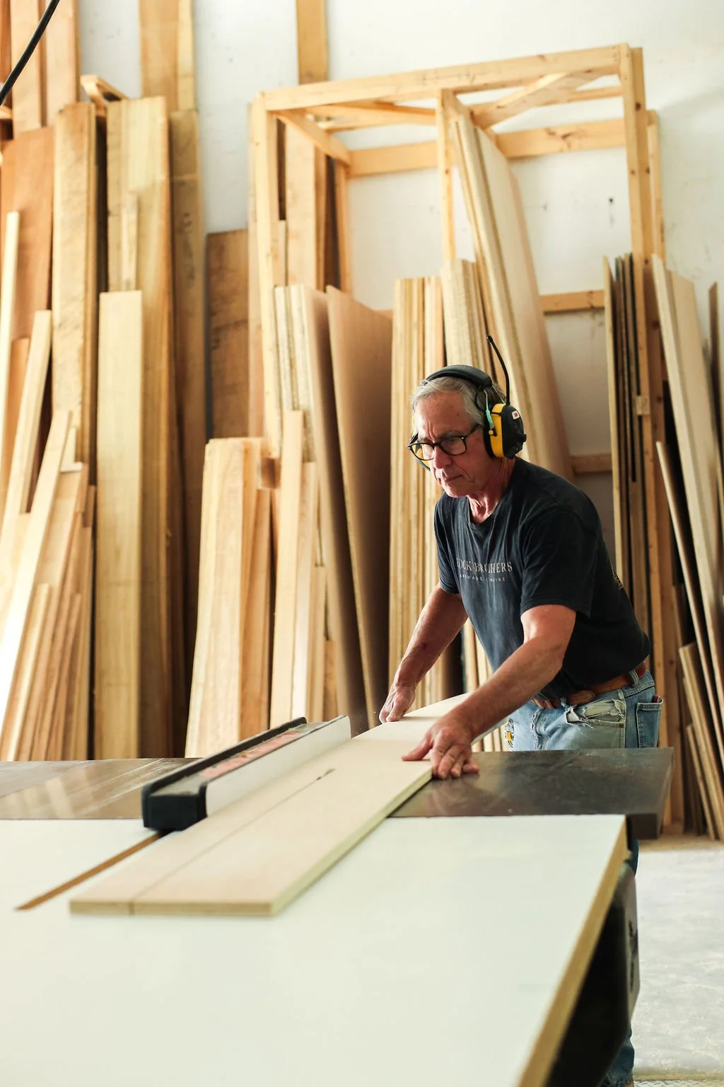 A man wearing safety ear protection and glasses working with a saw in a woodworking shop with various wooden boards stored on racks behind him.
