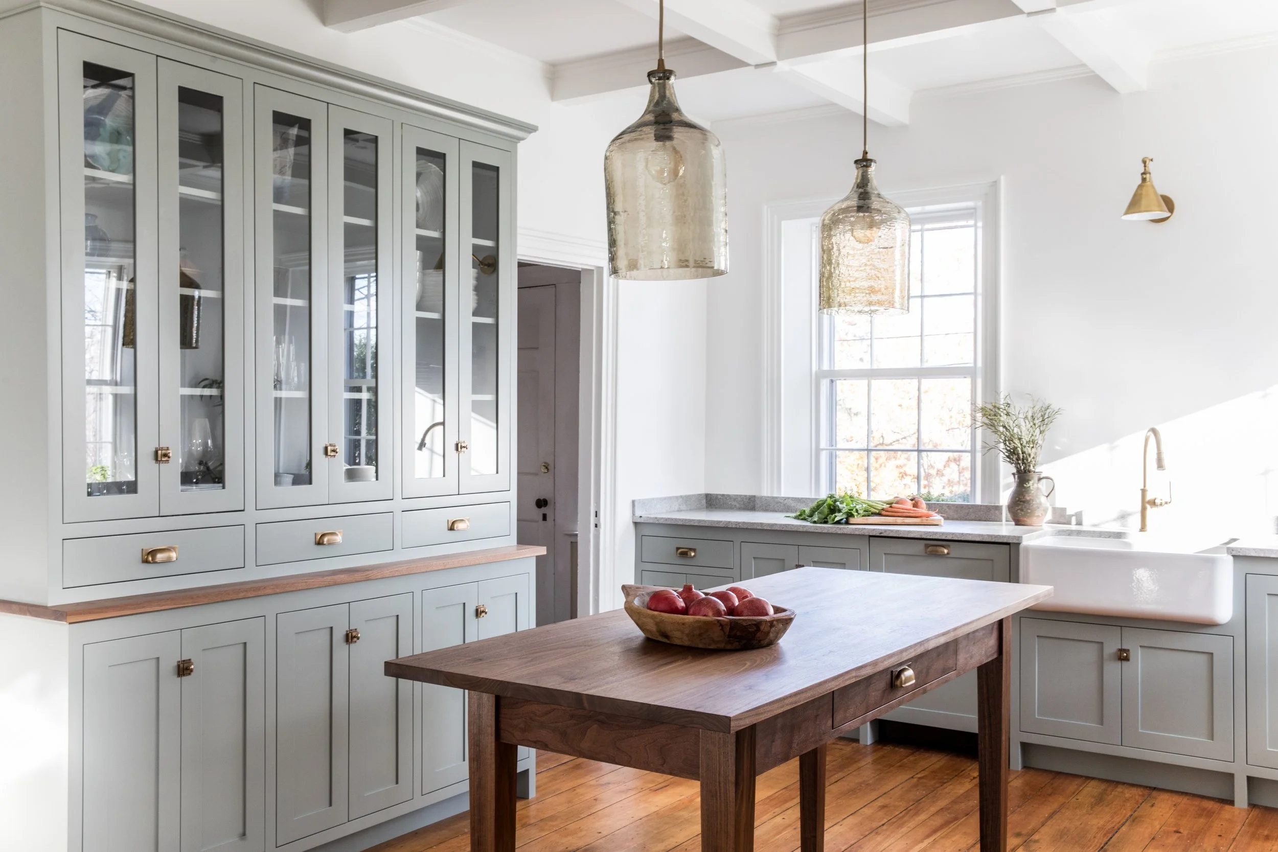 Bright kitchen with light gray cabinets, a white farmhouse sink, a wooden kitchen island with apples, large windows, hanging glass pendant lights, and a wooden floor.