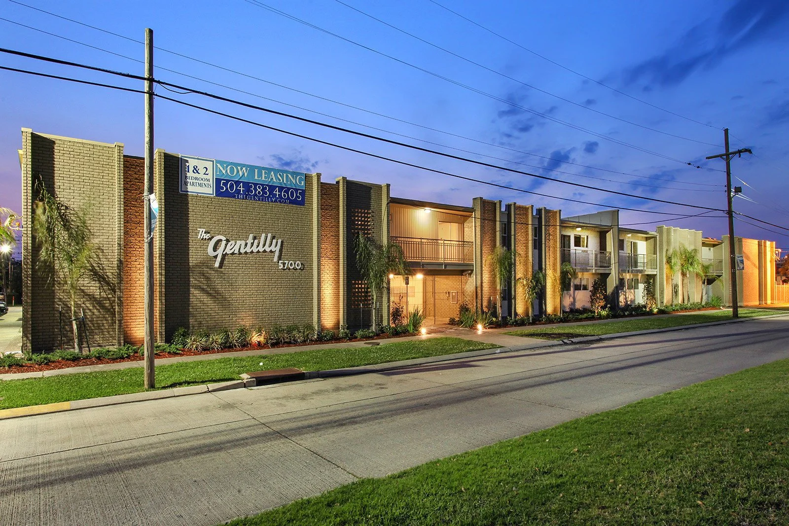 Exterior view of The Gently apartment complex at dusk, with lit building facade, palm trees, and a street in the foreground.