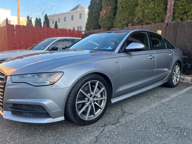 A silver sedan parked on a paved lot with a red wooden fence and trees in the background.