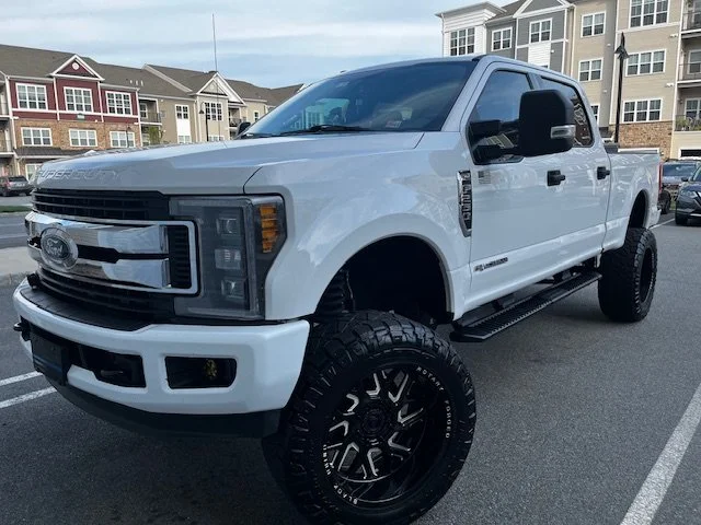 White Ford pickup truck with large off-road tires and black rims parked in a lot with apartment buildings in the background.
