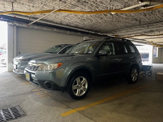 A black SUV parked in an indoor parking garage next to a silver sedan. The ceiling above has exposed pipes and wires.