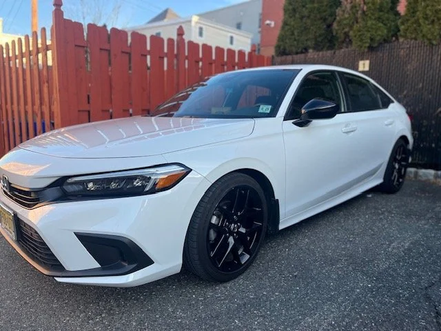 White sedan car with black wheels parked on asphalt next to a red wooden fence.