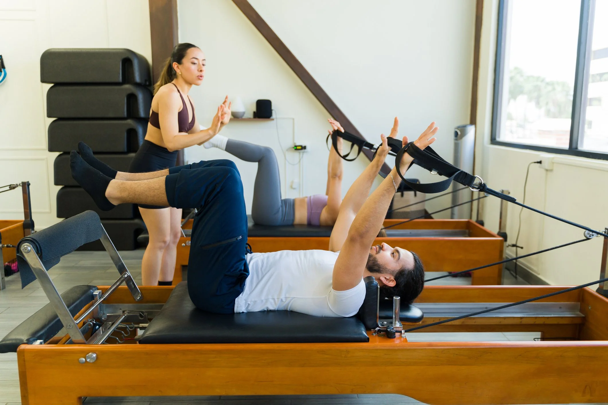 A person doing Pilates exercises on a Reformer machine guided by an instructor in a fitness studio.