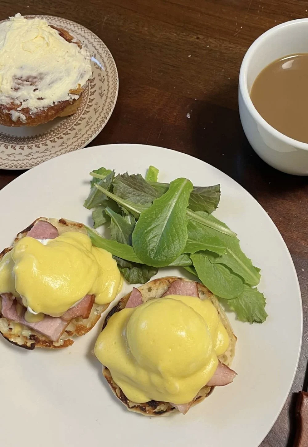 Plate with two eggs Benedict topped with hollandaise sauce, fresh green salad, a small dessert with icing and lemon icing, and a cup of coffee with cream on a wooden table.