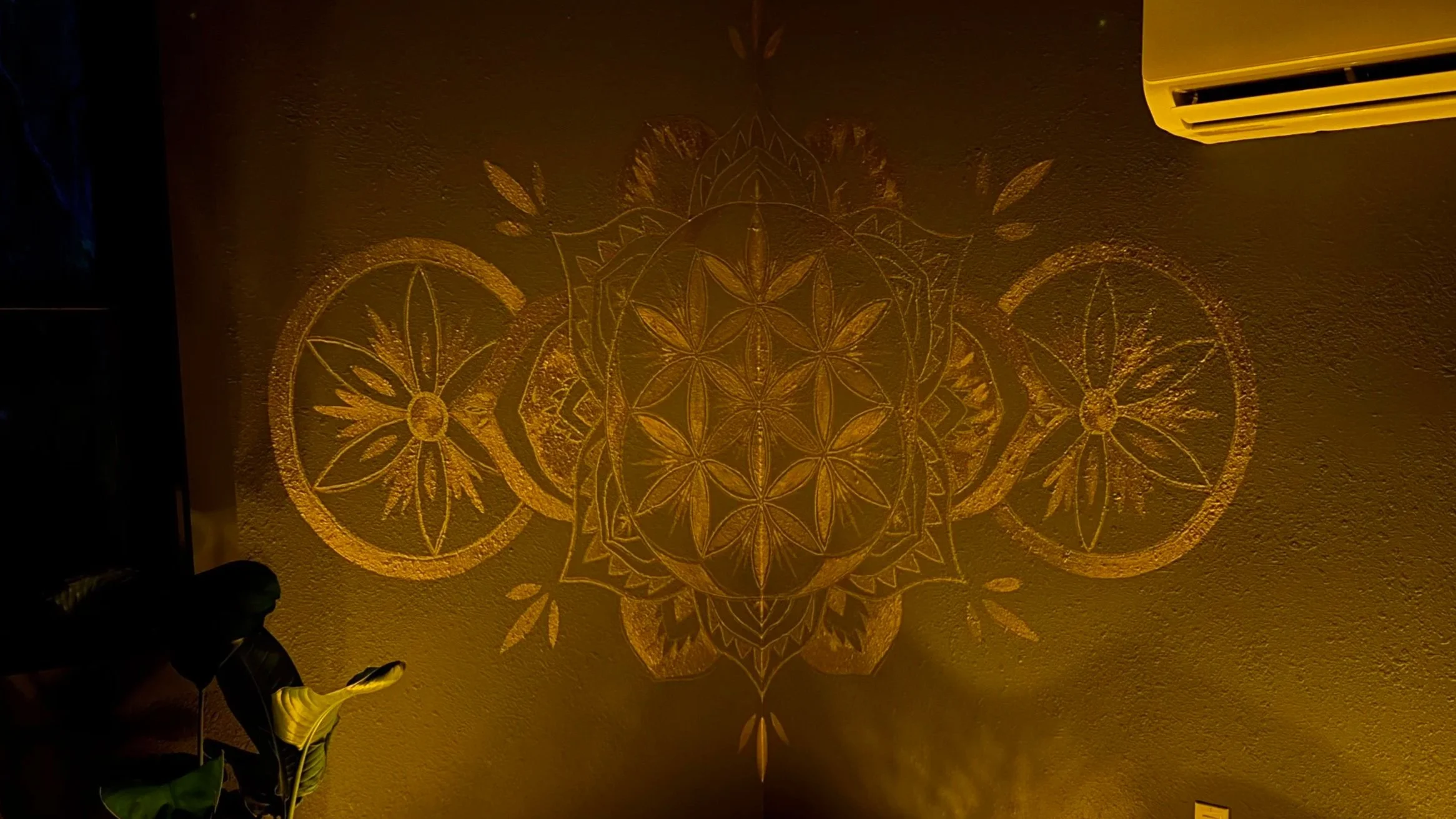 Intricate gold-colored geometric pattern projected onto a beige wall, illuminated by warm light, with a plant in the lower left corner and an air conditioning vent in the upper right corner.