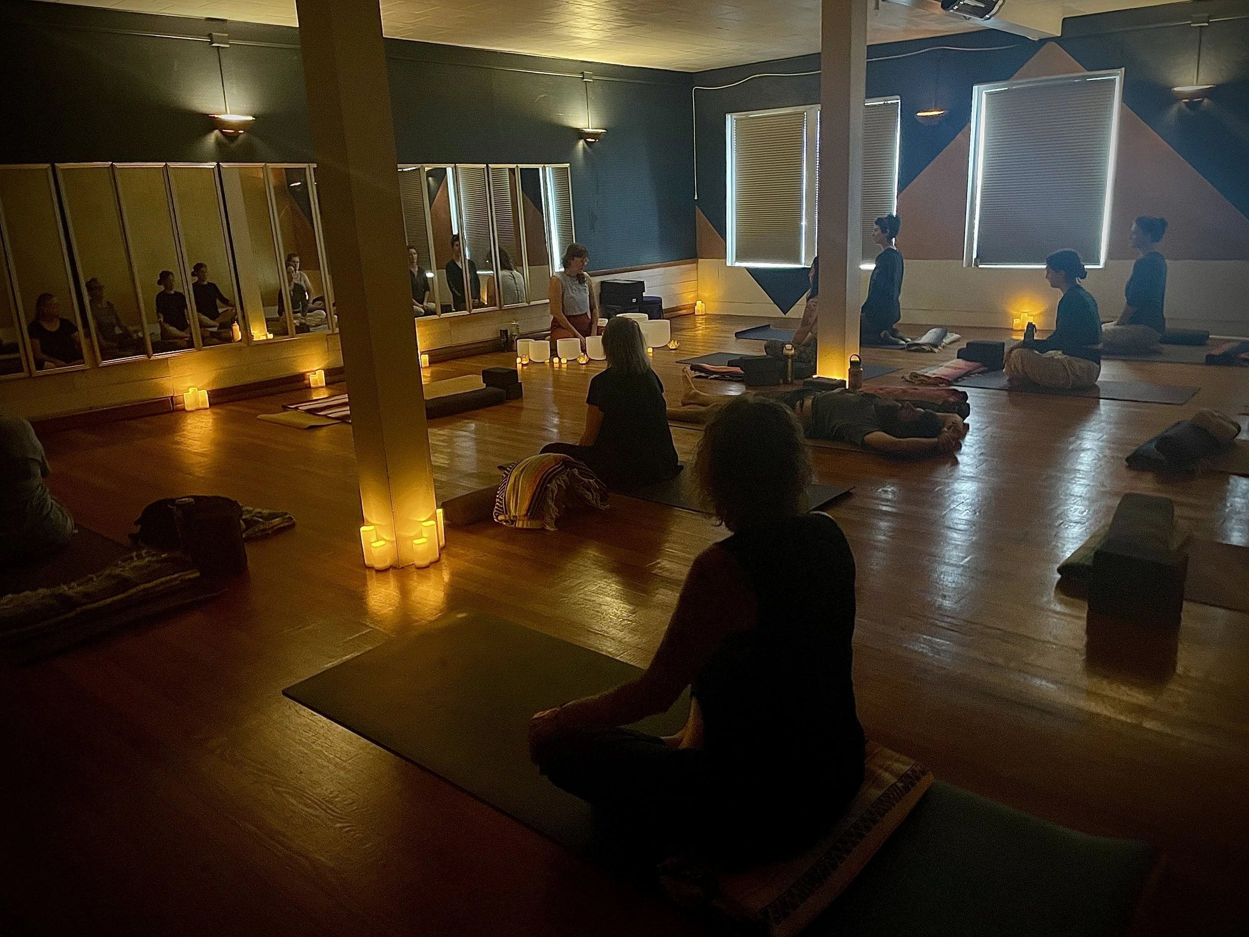 People participating in a meditation or yoga session in a dimly lit room with candles, windows with blinds, and a wooden floor.