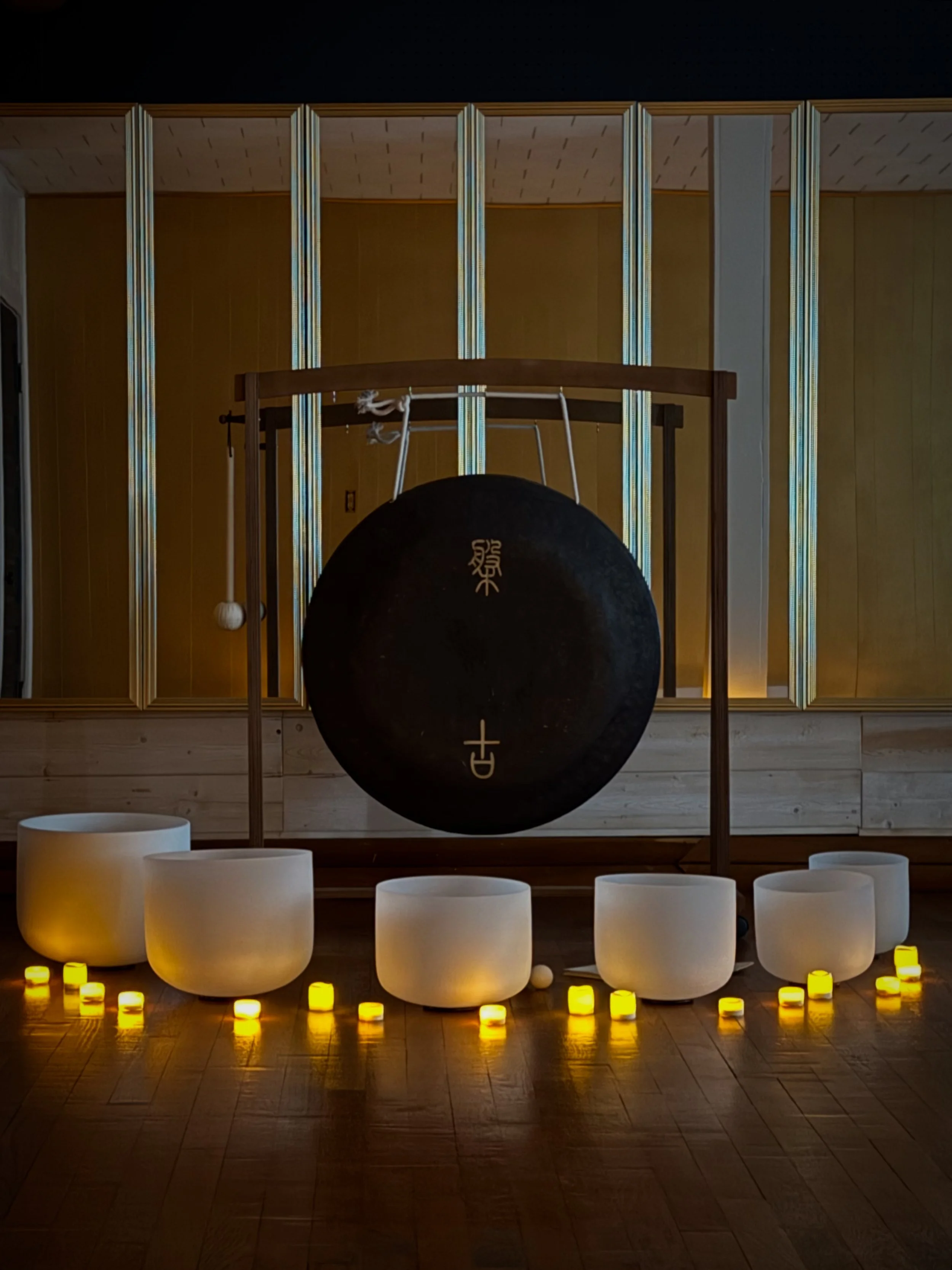 A musical setup with large white crystal singing bowls arranged in a semi-circle, illuminated by yellow candles on a wooden floor, and musical notes on a black ball in the center, with a background of yellow wooden panels and a large gong hanging on a wooden stand.
