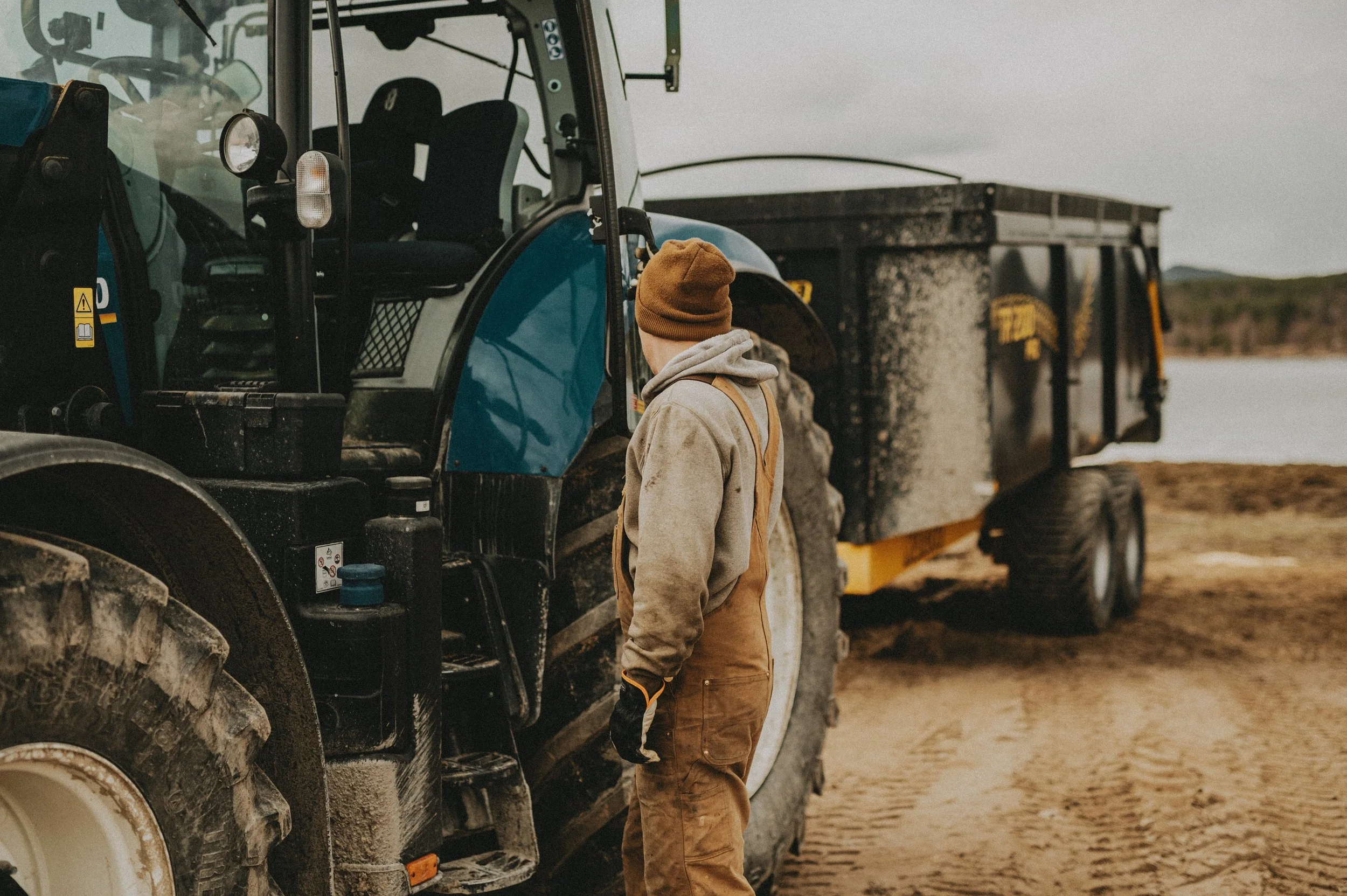 Person i arbetskläder stående bredvid en traktor vid en sjö, med utsikt över ett naturskönt landskap.