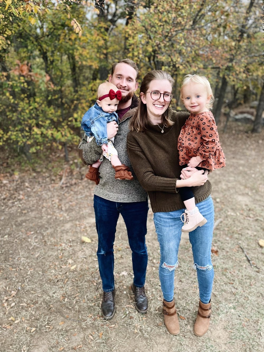 A smiling family of four outdoors in autumn, with fall foliage behind them. The father holds a baby girl with a red bow, the mother holds a toddler girl in a rust-colored dress. All are dressed casually.