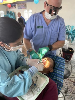 A dental team working on a young patient lying in a dental chair, with one dentist using tools inside the patient's mouth while the other stands nearby, in a dental clinic setting.