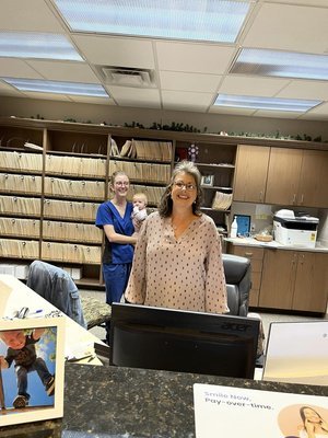 Two women and a girl standing behind a counter in an office or library, with shelves of files or magazines in the background.