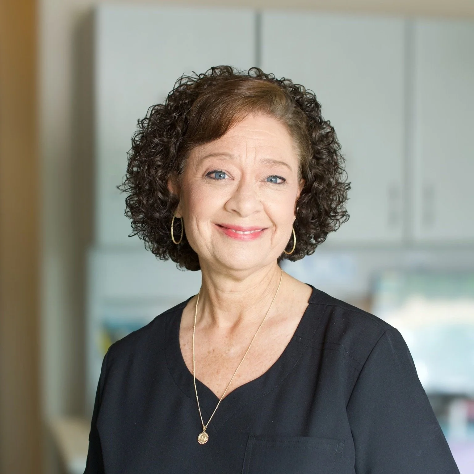 Smiling middle-aged woman with curly brown hair wearing a black top, gold hoop earrings, and a gold necklace, indoors with cabinets in the background.