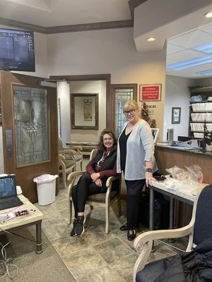 Two women in an office or waiting room, one seated in a chair and the other standing beside her, both smiling. There are glass doors, a TV screen, and shelves with files in the background.