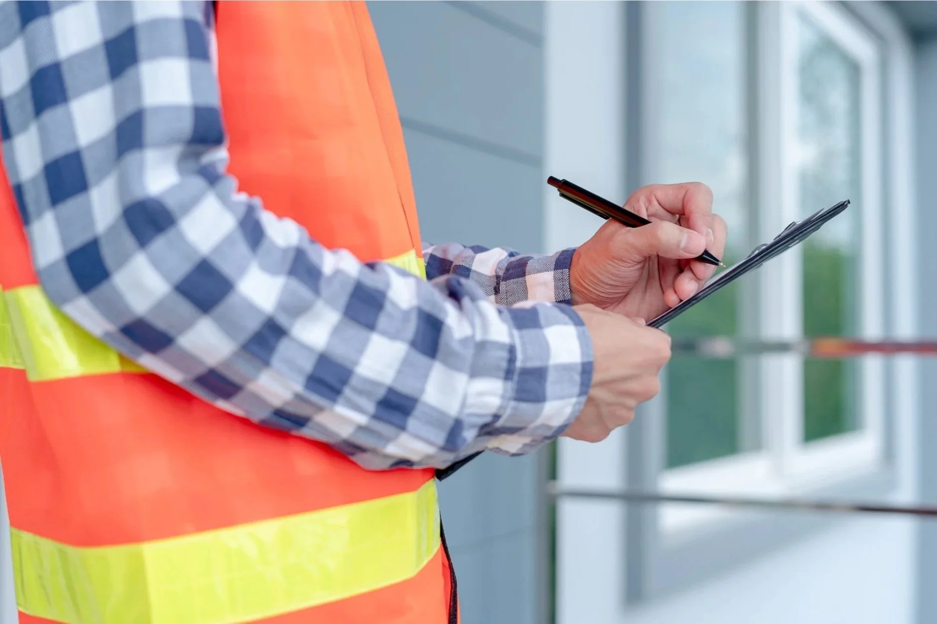 A WETT inspector worker wearing a high-visibility orange safety vest and blue checkered shirt, holding a WETT inspection report and a pen, standing outside a building.