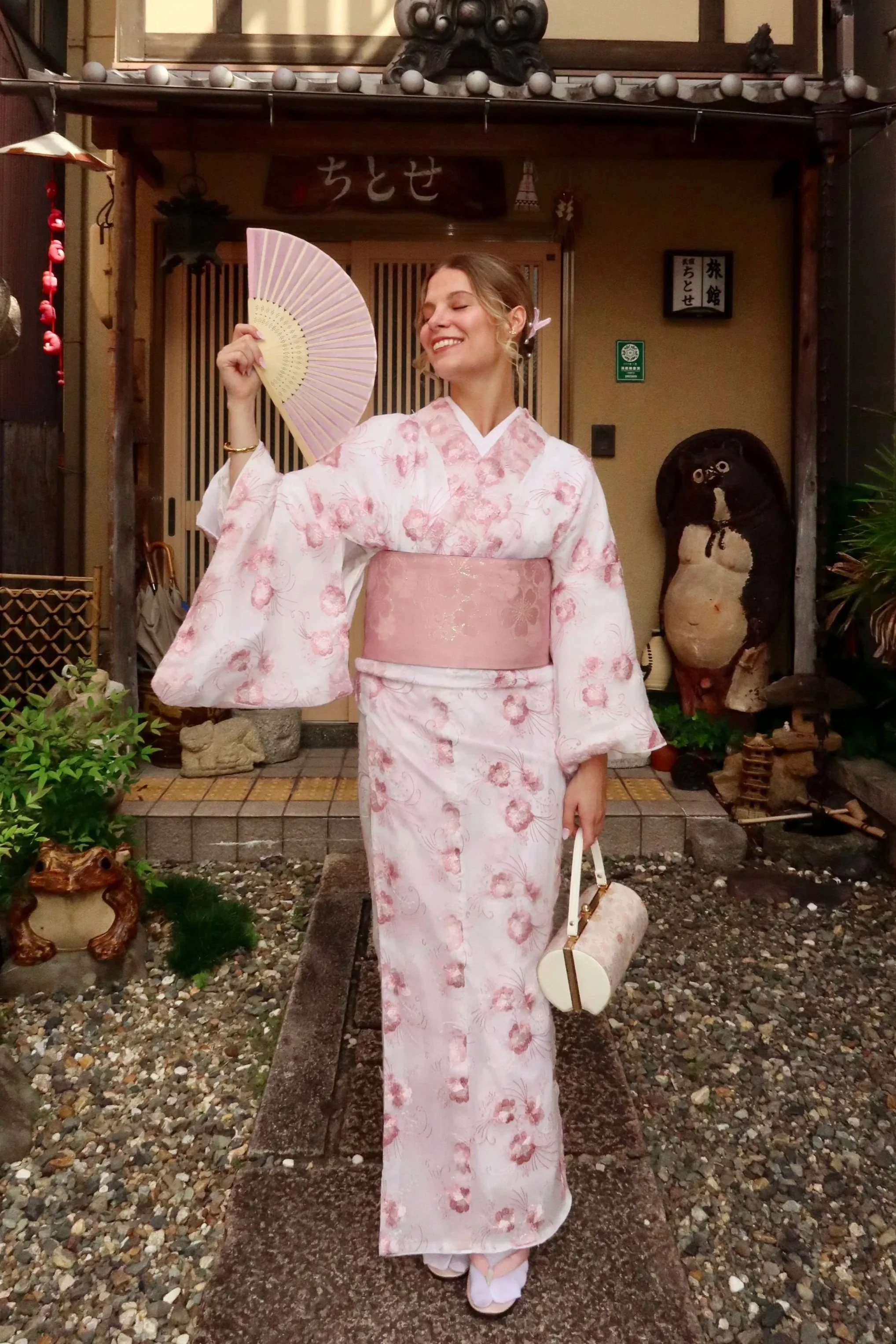 A woman in a pink floral kimono holding a folding fan and a small handbag, standing on a stone path outside a traditional Japanese building, smiling with her eyes closed.