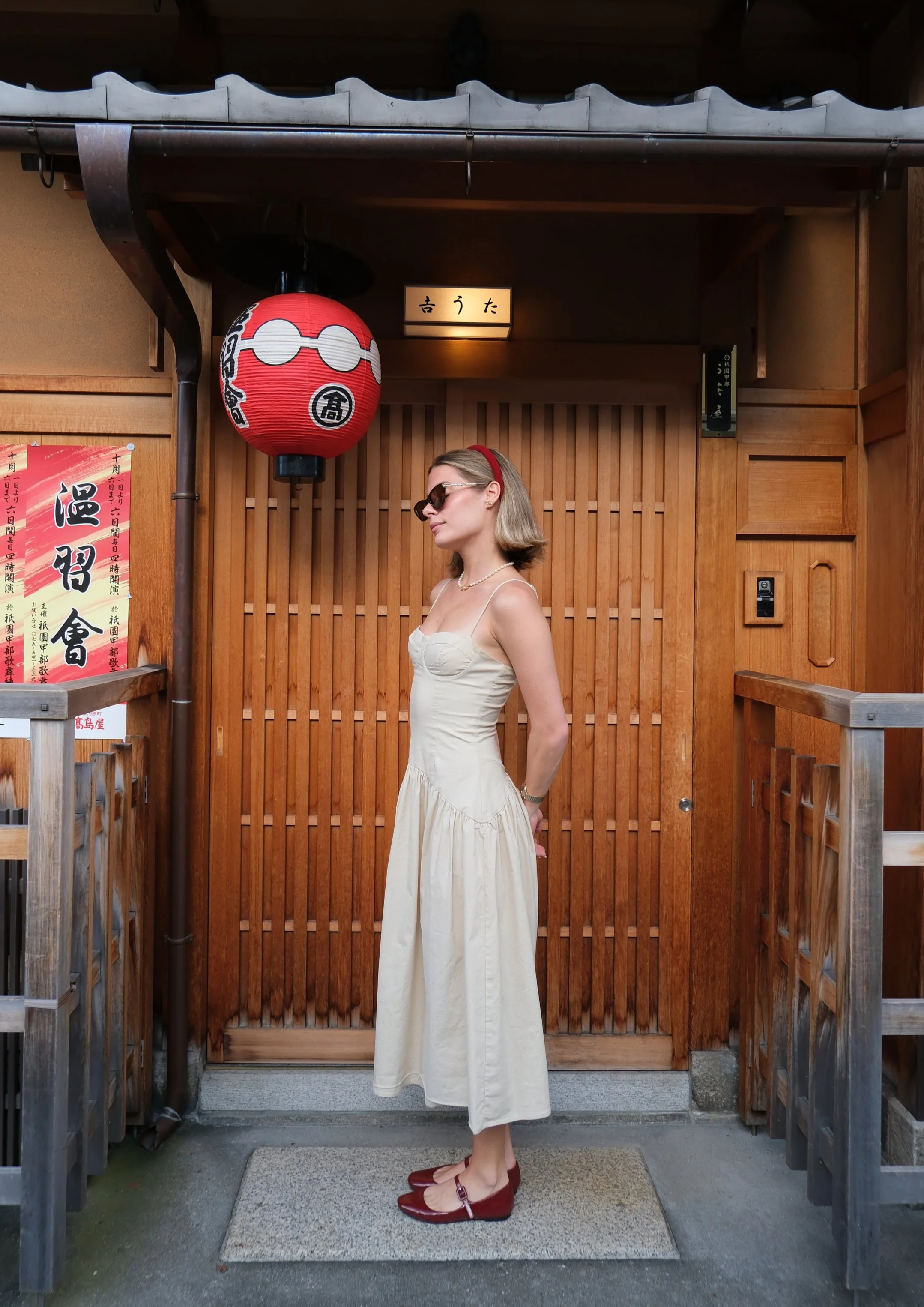 A woman with blonde hair wearing sunglasses, a headband, a white strapless dress, and red shoes standing in front of a wooden door with a red Japanese lantern hanging beside her.