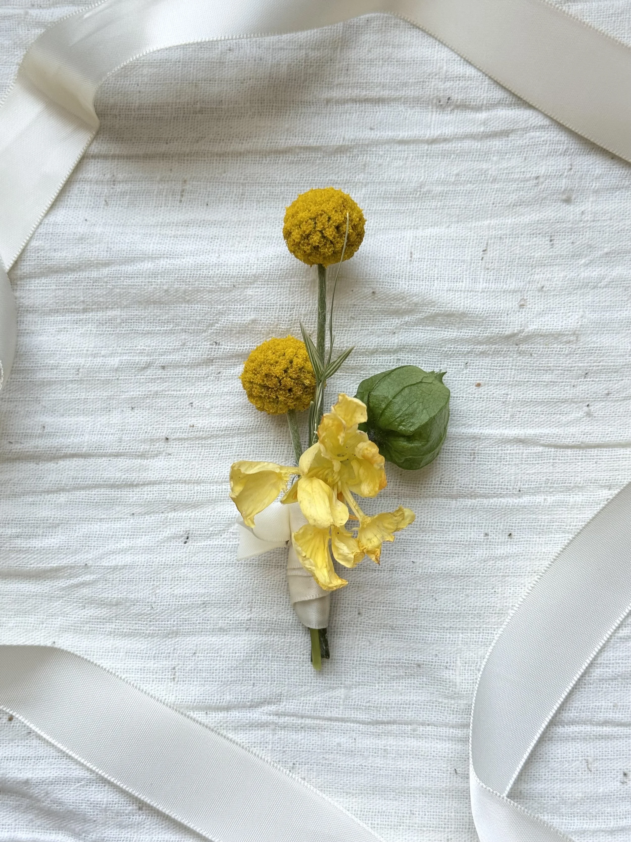 Yellow boutonniere with two round yellow flowers, a yellow iris, and a green leaf, pinned with a white ribbon on white textured fabric.